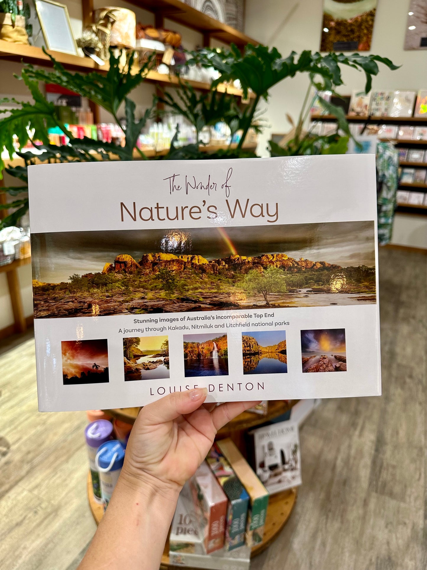 A hand holding a hard-cover coffee table book titled 'The Wonder of Nature's Way' by Louise Denton, showcasing photography from the Top End of Australia.